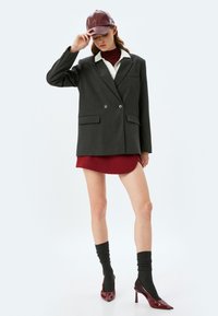 Grauer zweireihiger Blazer mit zwei Knöpfen, weißem Kragen, kombiniert mit einem kurzen bordeauxroten Kleid, dunklen Socken und bordeauxroten Lacklederspitzen.