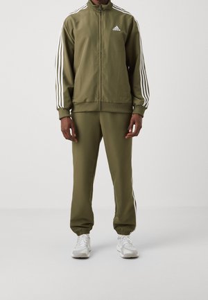 Personne portant un survêtement Adidas vert olive avec des bandes blanches sur les manches et le pantalon, assorti à des baskets blanches, debout devant un fond uni.