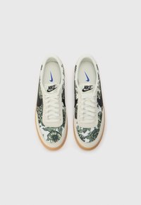 Un paio di scarpe Nike con un motivo floreale verde, logo swoosh nero, lacci bianchi, rifiniture beige e suola in gomma su sfondo bianco.