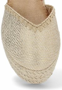 Alpargata beige con parte superior de textil tejido, que presenta una suela de yute trenzada y costuras decorativas a lo largo del borde. Diseño de punta redondeada y plana.