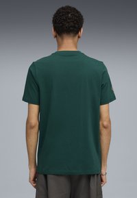 Ung person med kort krøllet hår iført en ensfarvet mørkegrøn t-shirt med korte ærmer og mørkegrå shorts, stående med ryggen til.