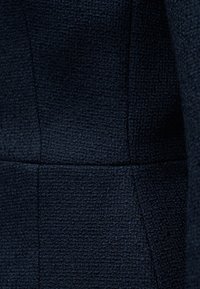 Κοντινό πλάνο σκούρου ναυτικού υφάσματος με υφή, με λεπτό ανάγλυφο τετράγωνο μοτίβο και ορατές ραφές.