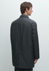 Schwarzer, langärmeliger Mantel mit geradem Schnitt und seitlichen Schlitzen. Hergestellt aus glattem, leichtem Stoff. Kragen und minimale Nähdetails sichtbar.