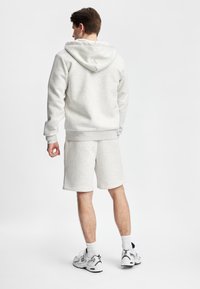 Mann iført lys grå hettegenser, matchende shorts, hvite sokker og hvite New Balance-sneakers som står med ryggen til på hvit bakgrunn.