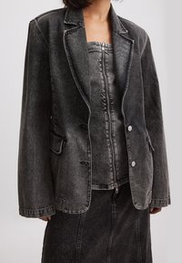 Giacca di jeans grigio con rever a punta, chiusura a doppio bottone, tasche frontali e cuciture a contrasto, indossata sopra un top coordinato con zip.