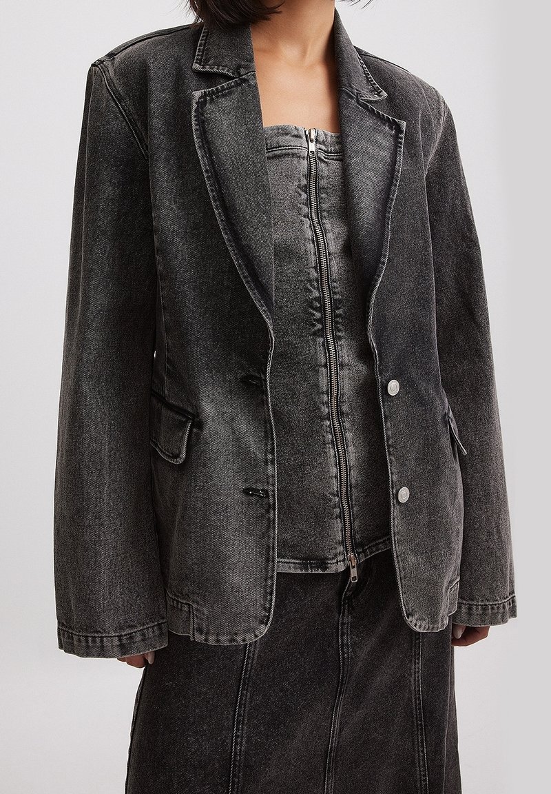 Giacca di jeans grigio con rever a punta, chiusura a doppio bottone, tasche frontali e cuciture a contrasto, indossata sopra un top coordinato con zip.
