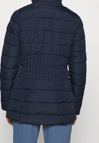 Veste matelassée bleu marine avec col haut, coutures horizontales et détails élastiques à la taille. Tissu lisse et coupe mi-longue.