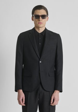 Joven hombre con traje negro, camisa negra y gafas de sol oscuras, de pie frente a un fondo liso de color gris claro.