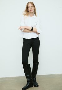 Weißes Hemd mit umgeschlagenen Ärmeln, kombiniert mit schwarzen Leggings und kniehohen schwarzen Stiefeln. Glattes Material und maßgeschneiderte Passform.