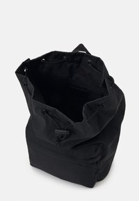 Sac à dos noir à cordon de serrage avec un extérieur en tissu doux, un fond structuré et une fermeture à cordon réglable. L'intérieur comporte une poche zippée sur le côté.
