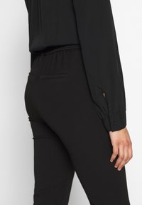 Mustat joustovyötärölliset housut, joissa virtaviivainen istuvuus. Takana tasku ja sileä kangas. Puserossa pitkät hihat ja napilliset kalvosimet.