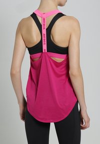 Femme portant un brassard de sport noir et un débardeur rose ample avec une lanière "JUST DO IT." dans le dos, debout devant un fond gris clair uni.