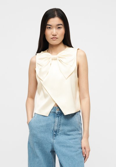 Γυναίκα με μακριά μαύρα μαλλιά, φορεί ένα αμάνικο κρεμ τοπ με μεγάλο φιόγκο και ανοιχτό μπλε τζιν ψηλόμεσο, με τα χέρια στις τσέπες.