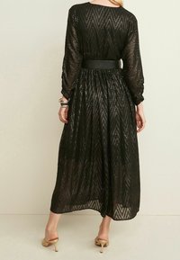 Vrouw in een zwarte doorschijnende langemouw jurk met een zigzagpatroon, een tailleband, gouden armbanden en beige hoge hakken, gezien van achteren.