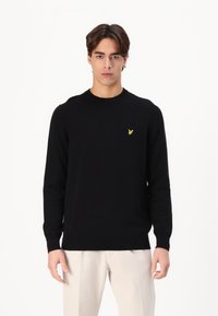 Zwart gebreid vest met een ronde hals, normale pasvorm, geribbelde manchetten en een klein geel logo op de borst. Gedomineerd met lichtgekleurde broeken.