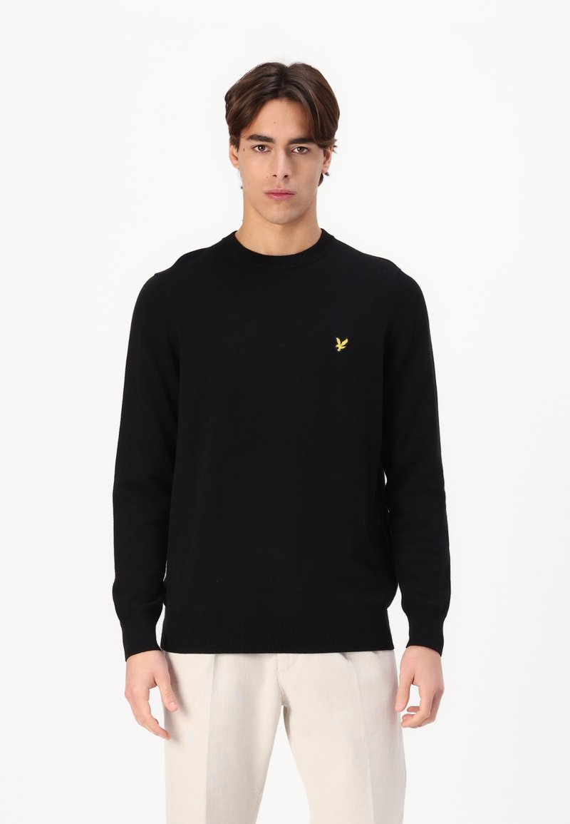 Zwart gebreid vest met een ronde hals, normale pasvorm, geribbelde manchetten en een klein geel logo op de borst. Gedomineerd met lichtgekleurde broeken.