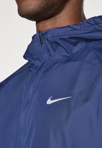 Navy Nike jacket with a lightweight, smooth texture; features a high collar, zipper, and a white logo accent on the front.