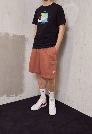 Osoba ubrana w czarną koszulkę Nike z nadrukiem, rdzawe szorty cargo Nike, białe skarpetki Nike oraz białe wysokie trampki, stojąca w pomieszczeniu.