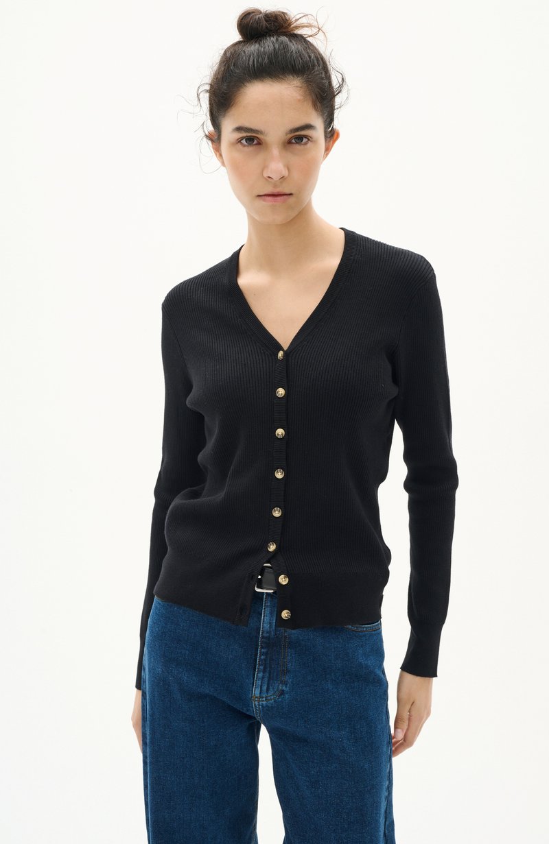 Svart ribbad cardigan med V-ringning, med guldknappar längs framsidan. Tillsammans med blå denimbyxor som framhäver en åtsittande siluett.