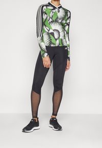 Långärmad, grön och svart mönstrad idrottströja med fågelmotiv, tillsammans med svarta leggings med nätpaneler och svarta sportskor.