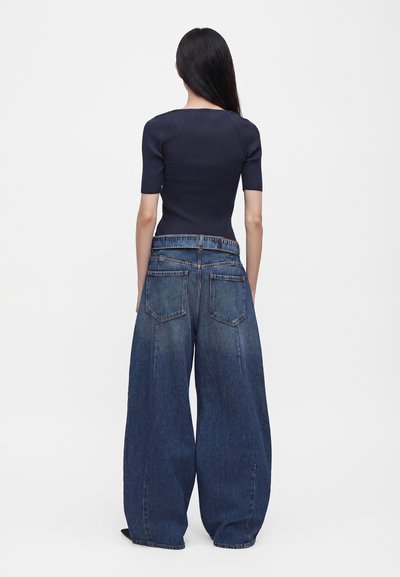 Γυναίκα με μακριά σκούρα μαλλιά φοράει εφαρμοστή μαύρη μπλούζα και φαρδιά μπλε τζιν, στέκεται με την πλάτη σε απλό ανοιχτό φόντο.