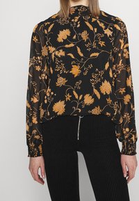 Blouse noire à fleurs avec des manches longues, présentant des motifs floraux orange et un col montant. Assortie à un pantalon côtelé noir.