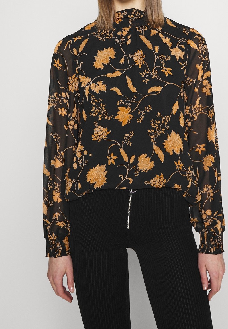 Blouse noire à fleurs avec des manches longues, présentant des motifs floraux orange et un col montant. Assortie à un pantalon côtelé noir.