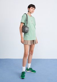 Mintgrønn t-skjorte med brodert design, beige plisserte skjørt, grå crossover-veske og grønne joggesko med hvite detaljer.
