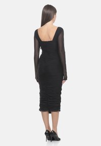 Robe noire avec des détails froncés, manches longues en dentelle, et un dos en V profond. Le tissu est lisse avec une silhouette ajustée, atteignant le mollet.