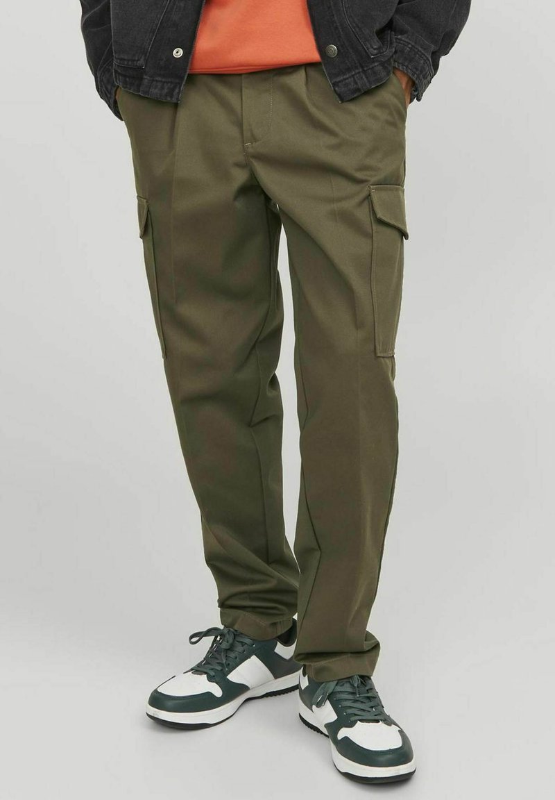Olivgrüne Cargo-Hosen mit Seitentaschen, schmalem Schnitt und sichtbaren Nähten. Kombiniert mit grünen und weißen Sneakers und einem orangefarbenen Shirt.