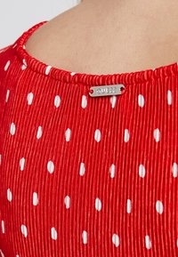 Tissu rouge à côtes avec des pois blancs, comportant une étiquette portant l'inscription "QUEEN" attachée au col. Texture lisse avec un léger éclat.