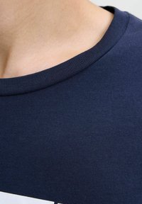 Sötétkék pamut póló kerek nyakkal és bordázott gallérral. Simább textúrával rendelkezik, és látható varrásokkal az éleken.