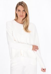 Valkoinen neulecardigan, jossa on napitettava etuosa, pitkät hihat, ribbauskohoumat hihoissa ja rento istuvuus, valmistettu pehmeästä kankaasta. Koristeena on pyöreä pääntie.