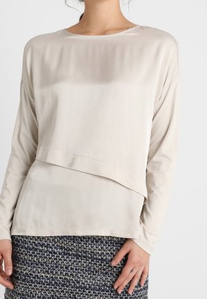Kobieta ubrana w luźną, beżową bluzkę z długim rękawem, z warstwowym detalem z przodu, oraz teksturowaną czarno-białą spódnicę.