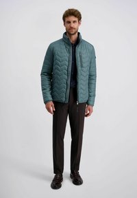 Man in een teal gewatteerde jas over een zwart shirt, donkere broek en zwarte nette schoenen, staand tegen een effen lichte achtergrond.