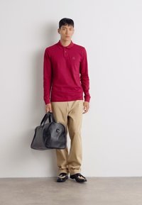 Bordeaux langsluitend poloshirt, beige broek, een zwarte leren tas dragend, zwarte schoenen met contrasterende details. Effen achtergrond.