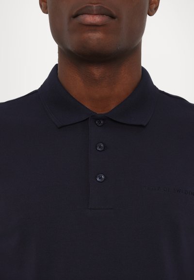 Tumesinine polo särk, mis on valmistatud tekstuuridest kangast, millel on volditud krae, kolme nupuga läbilöök ja rinnal diskreetne logopistik.