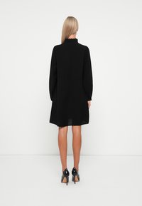 Svart langermet kjole med rynket krage, flytende design, kne-lang hem og båret med svarte stiletthæler. Glatt stoffstruktur.
