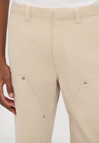 Pantalones beige con una textura suave, que cuentan con bolsillos inclinados asegurados con broches de metal y detalles cosidos a lo largo de la cinturilla.