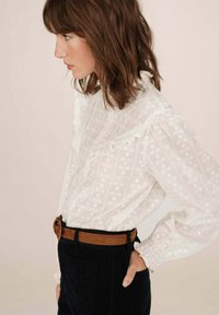 Blusa bianca con motivo testurizzato e colletto a volant, abbinata a pantaloni neri in corduroy e una cintura marrone. Vista di profilo.