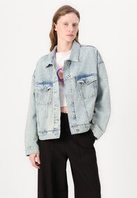 Light blue denim jacket with two chest pockets, buttons, and a slightly oversized fit. Features subtle patterned text throughout.