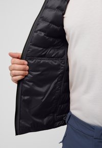 Schwarze gestepptenJacke mit Reißverschluss und gepolsterter Textur. Das Innere verfügt über eine Tasche und wird über einem hellen Hemd getragen.