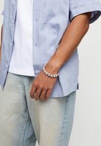 Camisa de manga corta azul claro con rayas verticales, combinada con jeans azul claro. Pulsera de cuentas plateadas en la muñeca, con superficie lisa y acabado reflectante.