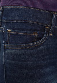 Los vaqueros de mezclilla azul oscuro presentan un bolsillo trasero distintivo, contorneado con costuras amarillas contrastantes y una textura suave.