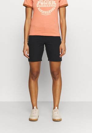 Femme portant un t-shirt corail avec le texte « Solar Power Regenerative Agriculture », un short noir au genou et des baskets beiges.