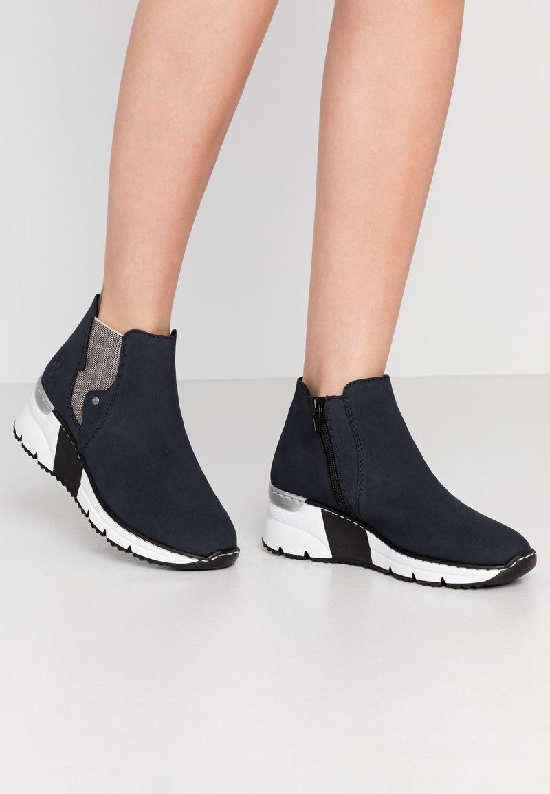 Baskets compensées bleu marine montant à la cheville avec fermetures éclair latérales et semelles à motifs blancs, noirs et argentés portées sur jambes nues.