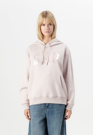 Lys rosa hettegenser laget av mykt materiale, med en frontlomme og shiny "GAP" bokstaver. Båret sammen med blå denimjeans.