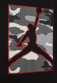 Černá tkanina s vyvýšenou grafikou zobrazující šedobílý maskovací vzor. Tučné červené šití rámuje černý siluet hráče.