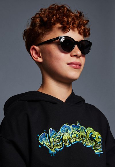 Juodas džemperis su ryškiu "Versace" grafiku mėlyna ir geltona blizgučiais. Asmuo dėvi juodus katės akių akinius ir turi garbanotus plaukus.