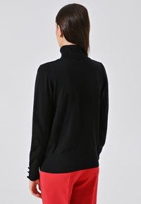 Frau mit langen dunklen Haaren, die einen schwarzen Rollkragenpullover und rote Hosen trägt, steht mit dem Rücken zu einem schlichten hellen Hintergrund.